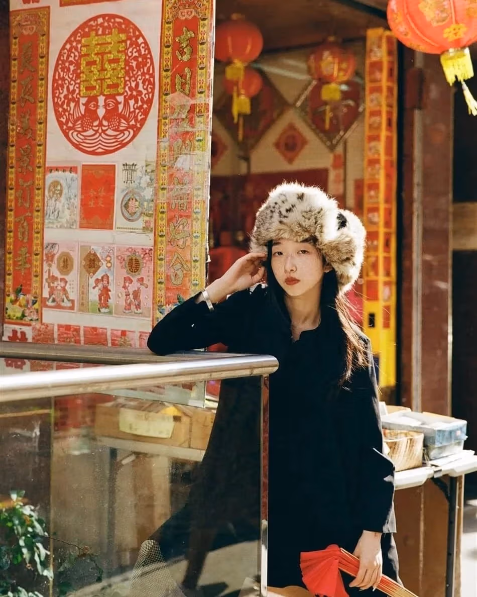 Image of a young woman with a big fuzzy hat, posing in front of a lot of traditional Chinese decorations..