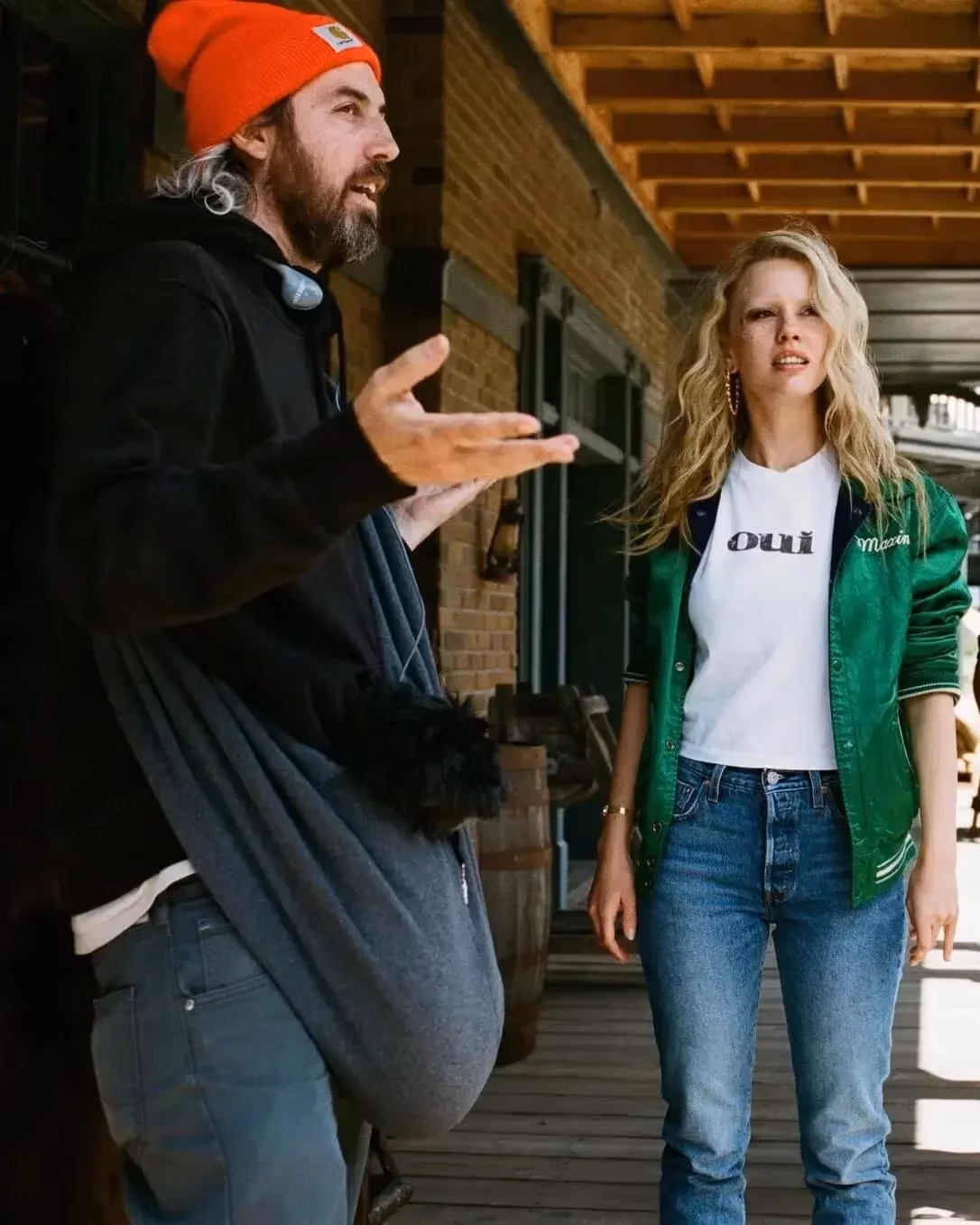 A man with long hair wearing a orange winter cap stands with his arms open, gesturing, with a blonde woman to his right wearing a bright green jacket and blue jeans.