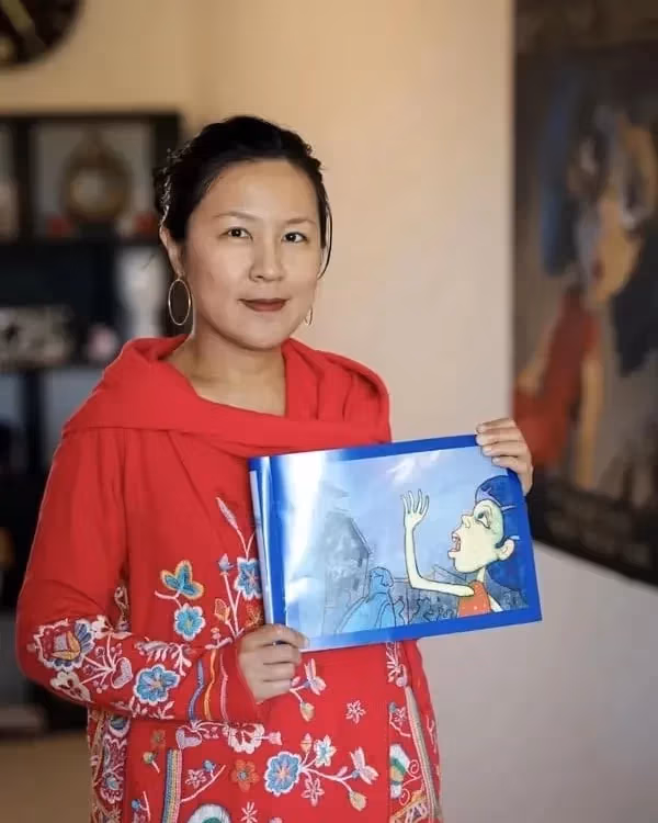 A woman in a red embroidered garment holds a children's book with an illustration of a shouting child. She stands indoors, conveying warmth and creativity.