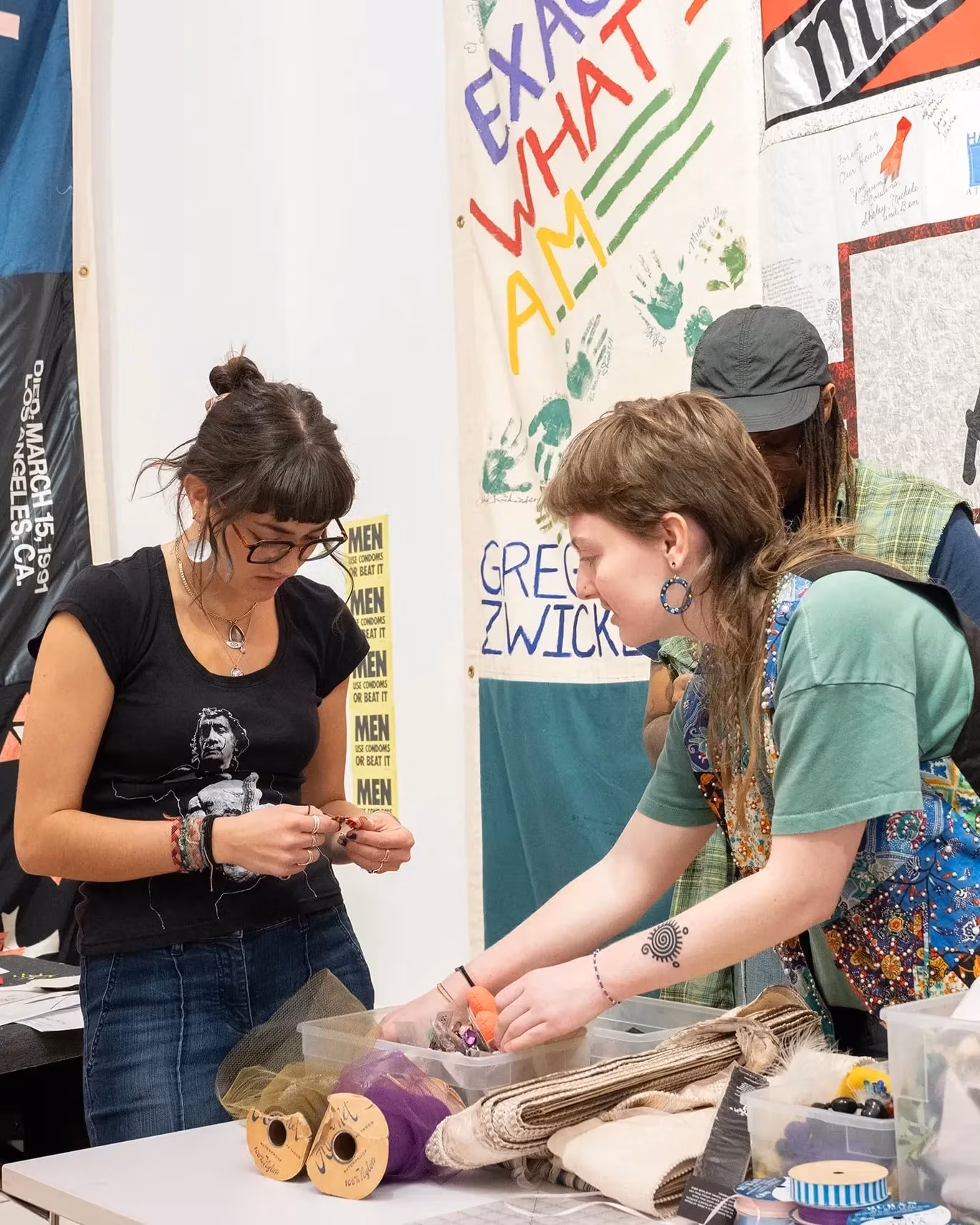 Three students are looking at fiber art materials in front of several printed artworks.